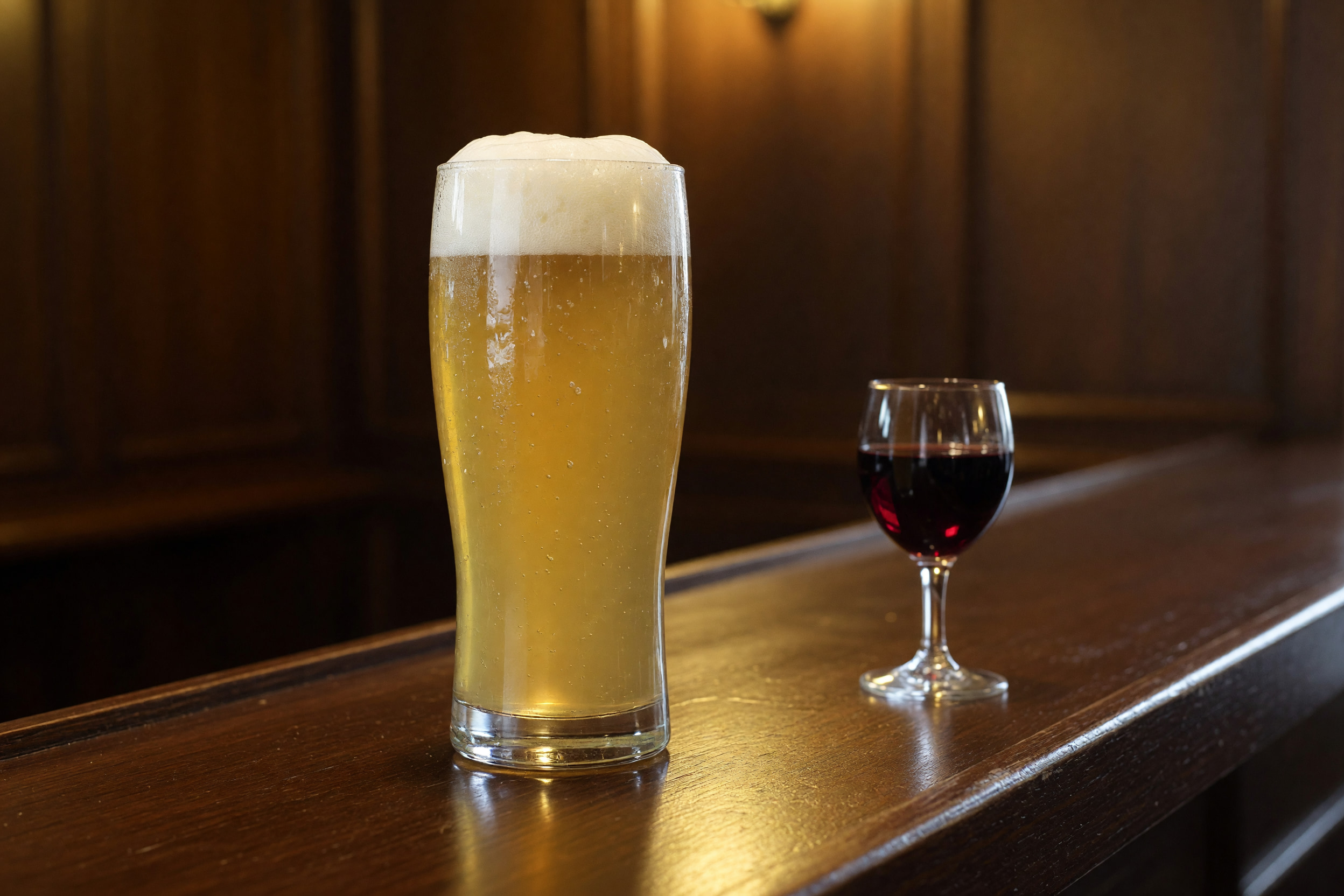 Glas naturtrübes Bier und Glas Rotwein auf dunkler Holztheke im warmen Wirtshauslicht